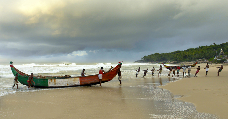pescatori tirano a riva una lunga barca  colorata su un distesa di sabbia contornata di palme